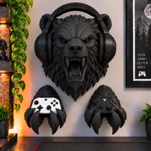 May include: A black wall-mounted bear head sculpture wearing black over-ear headphones, positioned above two black bear claw wall mounts, each holding a gaming controller, one white and one black, against a light wall.