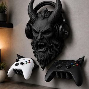 May include: A black wall-mounted display featuring a sculpted horned demon head with a beard, designed to hold gaming headphones. Below the head are two black claw-like wall mounts, each holding a wireless gaming controller, one white and one black.