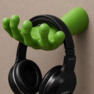 May include: A bright green wall-mounted hand sculpture designed as a headphone stand. The hand is positioned with fingers extended upwards to support a pair of black Edifier wireless over-ear headphones with cushioned ear cups and a headband.
