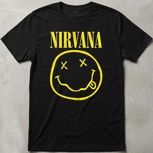 May include: A black short-sleeved t-shirt featuring the word NIRVANA in bold yellow capital letters above a yellow graphic of a smiley face with X-shaped eyes and a wavy mouth with a tongue sticking out, centered on the front of the garment.