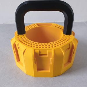 May include: A bright yellow, circular, multi-compartment tool organiser featuring a black handle. The top surface has a perforated grid pattern, and the sides contain several vertical slots designed for holding various small tools or accessories.