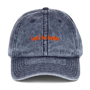 Puede incluir: Una gorra de béisbol azul oscuro con efecto lavado al ácido que presenta el texto naranja bordado Let's be better centrado en el panel frontal. La gorra tiene una visera curva, un botón en la parte superior y ojales cosidos para ventilación.