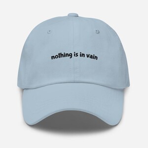 May include: A light blue baseball cap featuring the embroidered black text "nothing is in vain" centred on the front panel. The hat has a structured crown, a curved brim, and a button at the top, showcasing a clean and simple design.