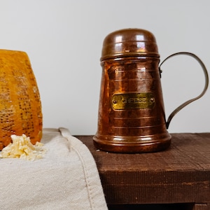 May include: A copper cheese shaker with a rounded lid and a curved handle. A rectangular brass plaque on the front is engraved with the word CHEESE. The shaker is positioned next to a wedge of hard cheese and a small pile of grated cheese on a cloth.