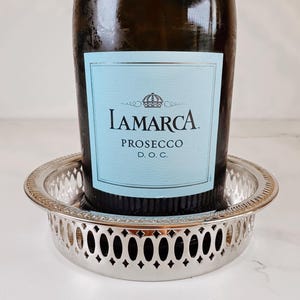 May include: A dark glass bottle of La Marca Prosecco D.O.C. with a light blue label featuring a crown icon, resting inside a round, silver-toned metal coaster with a decorative pierced lattice pattern along the vertical rim.
