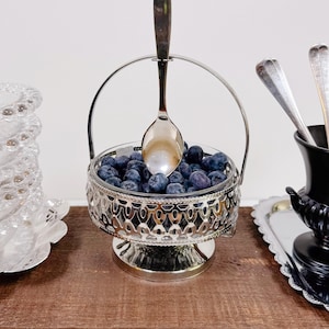 Könnte beinhalten: Eine silberfarbene Servierschale aus Metall mit dekorativem Filigranmuster und einem hohen, bogenförmigen Griff. Die Schale ist mit frischen Blaubeeren gefüllt und enthält einen silbernen Löffel, der aufrecht im Behälter steht.