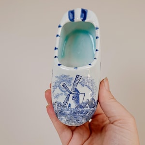 May include: A white ceramic clog-shaped ornament featuring a blue transferware-style print of a windmill and rural landscape. The rim is decorated with blue stripes, and the interior is glazed in a light blue hue, creating a decorative object.