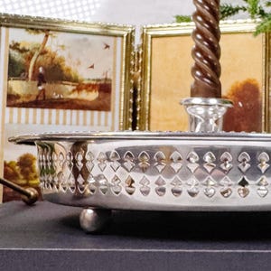 May include: A silver-toned metal tray with a pierced lattice pattern along the side and small ball feet. A single brown twisted taper candle stands in a central holder. The tray has a polished finish and a shallow, circular design.