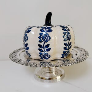 Puede incluir: Una figura de calabaza de cerámica con un acabado brillante de color crema y patrones de enredaderas florales azules pintados en sus nervaduras. La calabaza tiene un tallo negro sólido y se exhibe sobre un soporte de pedestal de metal plateado.