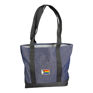 Op de afbeelding: Een donkerblauwe denim tote bag met twee zwarte stoffen hengsels en een zwarte verstevigde onderkant. Een rechthoekige patch met de Progress Pride vlag, voorzien van horizontale regenboogstrepen en een chevron-ontwerp, is in het midden geplaatst.