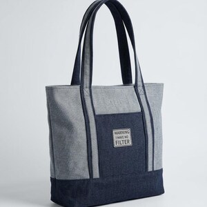 May include: A two-tone tote bag featuring light grey and dark navy blue fabric panels. The bag has two long navy blue shoulder straps and a rectangular front pocket with a patch that reads WARNING I HAVE NO FILTER in black text.