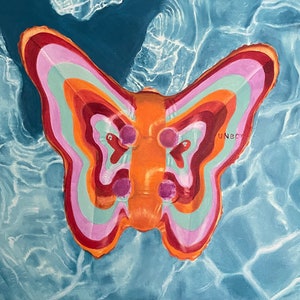 Puede incluir: Un flotador inflable con forma de mariposa en tonos naranja, rosa, rojo y turquesa. El flotador está en una piscina de agua azul con reflejos de luz. La palabra "UNBC" está impresa en el flotador.