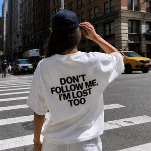 Op de afbeelding: Een wit T-shirt met korte mouwen en de zwarte tekst "DON'T FOLLOW ME, I'M LOST TOO" op de achterkant. Het shirt wordt gecombineerd met een donkerblauwe baseballpet en een witte broek, in een stedelijke omgeving met een gele taxi.