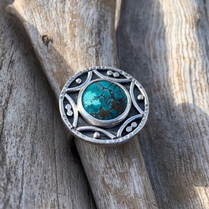 May include: A silver ring with a turquoise stone set in a circular design. The ring is on a wooden surface.