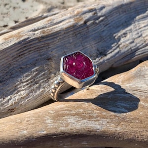 May include: A silver ring featuring a large, faceted, deep pink gemstone. The gemstone is set in a geometric bezel. The ring's band has a textured design. The ring is displayed on a piece of driftwood.