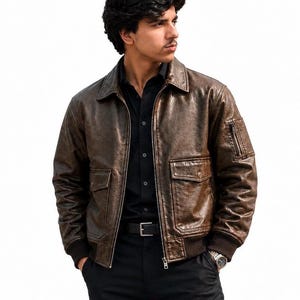 A dark brown leather bomber jacket featuring a distressed texture, a classic collar, a front zipper closure, two flap pockets at the waist, and a small zippered utility pocket on the left sleeve with ribbed cuffs and a ribbed hem.