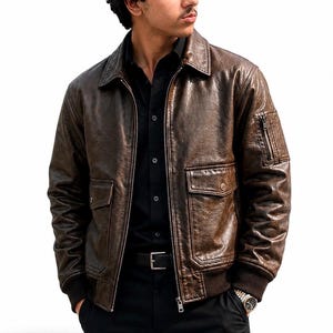 A dark brown leather bomber jacket featuring a distressed texture, a classic collar, a front zipper closure, two flap pockets at the waist, and a small zippered utility pocket on the left sleeve with ribbed cuffs and a ribbed hem.