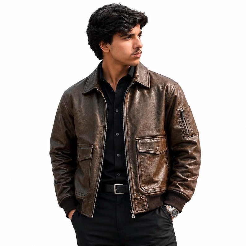 A dark brown leather bomber jacket featuring a distressed texture, a classic collar, a front zipper closure, two flap pockets at the waist, and a small zippered utility pocket on the left sleeve with ribbed cuffs and a ribbed hem.
