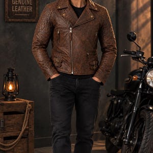 Pu&ograve; includere: Una giacca da moto in pelle marrone con motivo trapuntato sulle spalle, chiusura diagonale con zip argentata, due tasche laterali con zip e fibbie laterali regolabili in vita, indossata sopra una maglietta nera e jeans scuri.