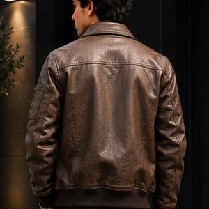 A brown leather bomber jacket featuring a textured finish, a classic collar, long sleeves with a small zippered pocket on the upper arm, and a ribbed hem and cuffs. The jacket is worn over dark trousers, showcasing a structured fit.
