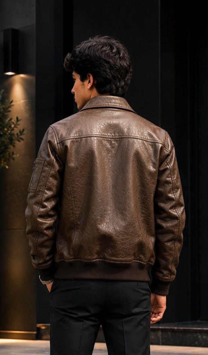 A brown leather bomber jacket featuring a textured finish, a classic collar, long sleeves with a small zippered pocket on the upper arm, and a ribbed hem and cuffs. The jacket is worn over dark trousers, showcasing a structured fit.