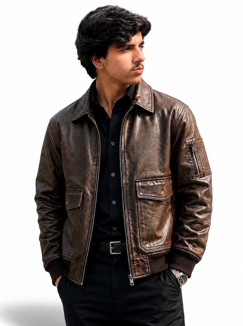 A dark brown leather bomber jacket featuring a distressed texture, a classic collar, a front zipper closure, two flap pockets at the waist, and a small zippered utility pocket on the left sleeve with ribbed cuffs and a ribbed hem.