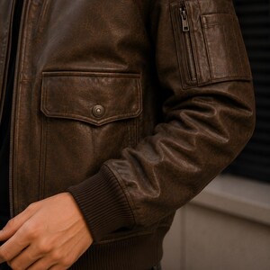 A dark brown leather bomber jacket featuring a front zipper closure, a snap-button flap pocket on the side, and a zippered utility pocket on the upper sleeve. The jacket includes dark brown ribbed cuffs and a matching ribbed hemline.