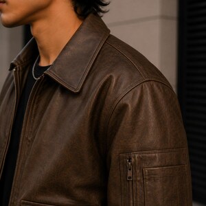 A dark brown leather jacket featuring a classic collar, a full-length front zipper, and a utility pocket on the upper sleeve with a vertical zipper closure. The jacket has a textured finish and is worn over a plain black shirt.