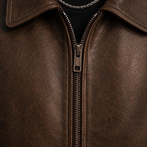 A dark brown leather jacket featuring a textured surface, a classic collar, and a central metal zipper closure. A thin silver-toned chain necklace is visible around the neckline, resting against a black base layer underneath the jacket.