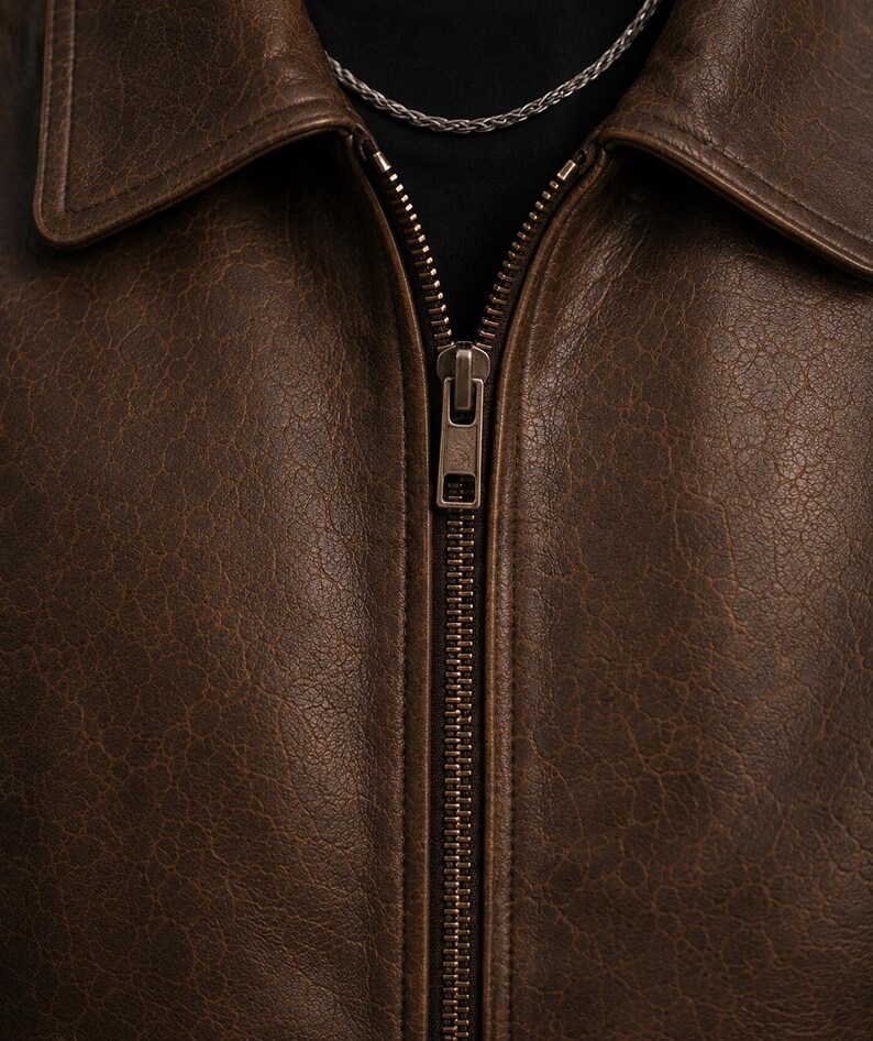 A dark brown leather jacket featuring a textured surface, a classic collar, and a central metal zipper closure. A thin silver-toned chain necklace is visible around the neckline, resting against a black base layer underneath the jacket.