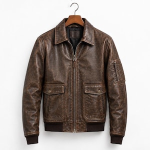 A brown leather bomber jacket featuring a front zipper closure, a classic collar, and two large flap pockets with snap buttons. The jacket includes a small zippered utility pocket on the left sleeve and ribbed cuffs and hem.