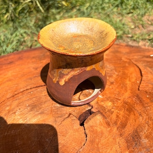 May include: A ceramic oil burner featuring a wide, shallow bowl top with a mottled yellow and orange glaze, transitioning into a dark brown base with a large, arched cutout opening for a tea light candle at the front of the vessel.