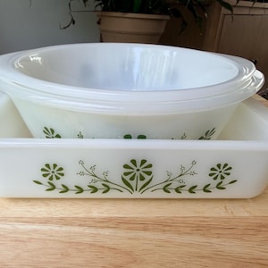 May include: A white milk glass square baking dish and a round mixing bowl, both featuring a green floral pattern of daisies and leafy stems. The square dish has a flat rim, while the round bowl has a wide, smooth edge.