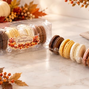 May include: A clear plastic container filled with chocolate, vanilla, and caramel macarons, featuring a label that reads Happy Thanksgiving. Next to the box, four additional macarons in brown, yellow, and cream colors are arranged in a tray.