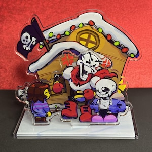 May include: An acrylic standee featuring three cartoon characters in front of a snow-covered wooden house with a pirate flag. One character is a tall skeleton with red gloves, another is a shorter skeleton in a blue jacket, and the third is a child.