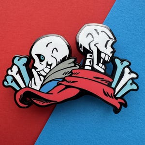 May include: A metal enamel pin featuring two cartoon skeletons side by side. One skeleton has a blue eye and a grey hoodie, while the other wears a long red scarf. The design is accented with light blue and white bone shapes on both outer edges.