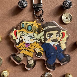 Könnte beinhalten: Ein Acryl-Schlüsselanhänger mit zwei Zeichentrickfiguren. Eine Figur trägt einen gelben Trenchcoat und eine Schutzbrille, die andere einen dunkelblauen Anzug und einen Fedora. Der Anhänger hat einen dunklen Metallkarabiner und ist von kleinen Metallperlen umgeben.