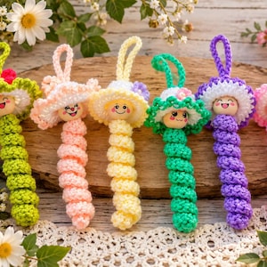 May include: Six handmade crochet ornaments featuring smiling wooden bead faces and wide-brimmed hats. Each ornament has a long, spiraled body in vibrant colors including lime green, pale pink, yellow, teal, purple, and light pink, with a hanging loop.