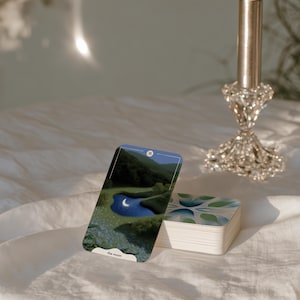 Könnte beinhalten: Eine Tarotkarte mit der Aufschrift the moon und der Nummer 18, die eine dunkle Landschaft mit einer Mondsichel in einem blauen Teich zeigt, gelehnt an einen Kartenstapel neben einer hohen, brennenden Metallkerze in einem verzierten Glashalter.