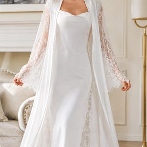 May include: A floor-length white satin slip dress with a cowl neckline, paired with a matching long-sleeved robe featuring intricate white lace trim on the sleeves and side panels. The ensemble is crafted from a smooth, solid white fabric.