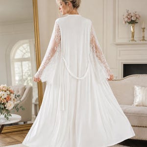 May include: A floor-length white robe featuring long, sheer lace sleeves with intricate floral patterns. The garment includes a matching white fabric tie belt at the waist, a smooth solid white back panel, and a flowing, loose-fitting silhouette.