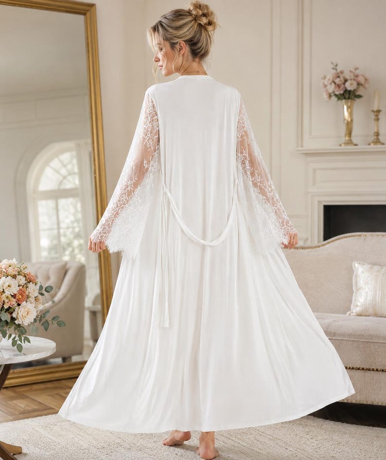 May include: A floor-length white robe featuring long, sheer lace sleeves with intricate floral patterns. The garment includes a matching white fabric tie belt at the waist, a smooth solid white back panel, and a flowing, loose-fitting silhouette.
