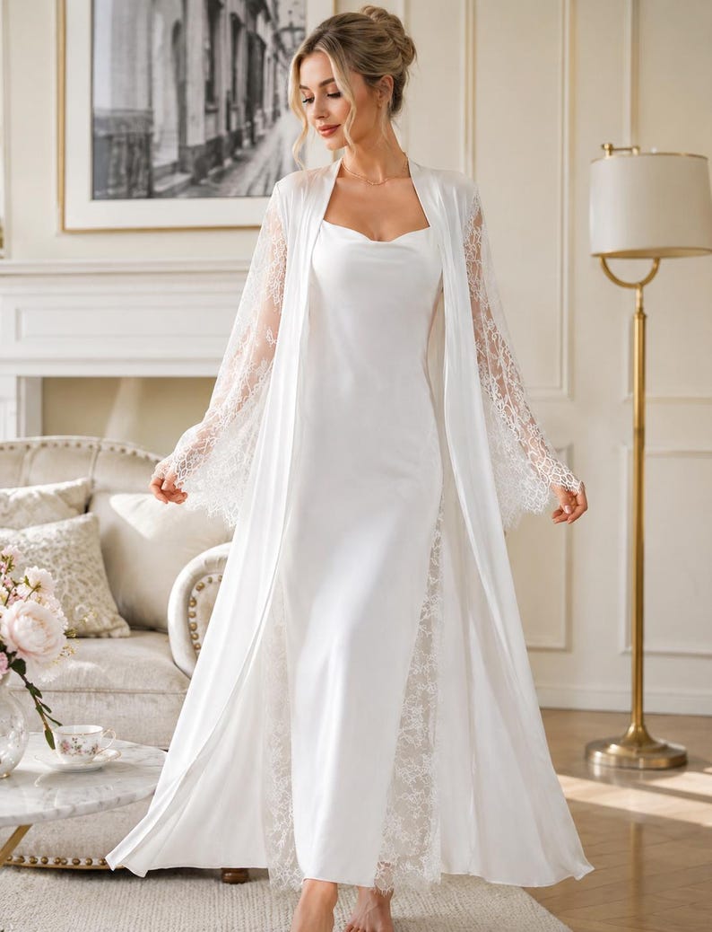 May include: A floor-length white satin slip dress with a cowl neckline, paired with a matching long-sleeved robe featuring intricate white lace trim on the sleeves and side panels. The ensemble is crafted from a smooth, solid white fabric.