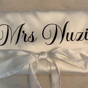 May include: A white satin robe featuring a tie-waist belt and the text Mrs Nuzi printed in a black cursive script across the front. The fabric has a smooth, reflective finish and is folded neatly with the sash tied in a bow at the center.