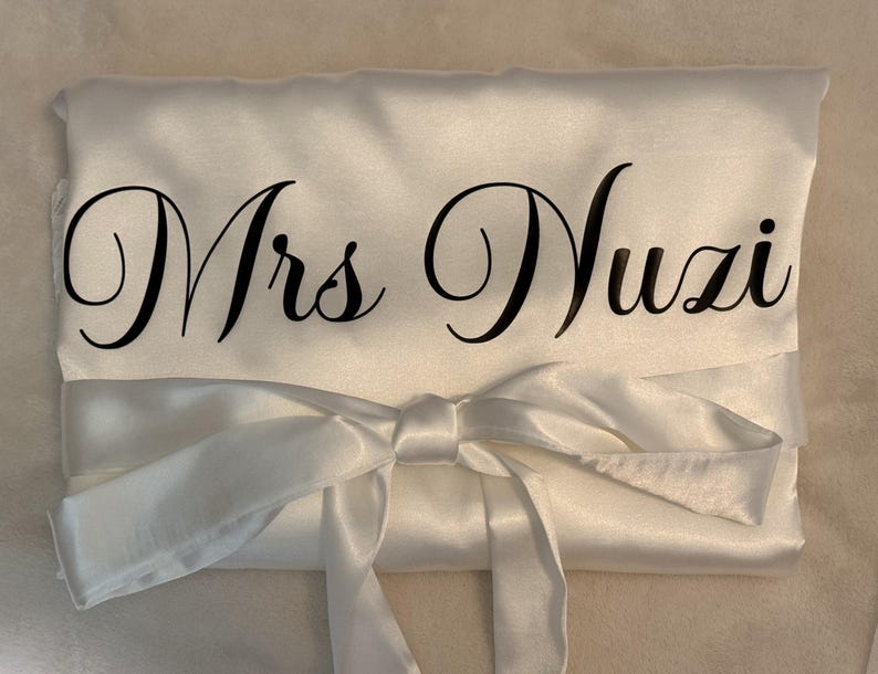 May include: A white satin robe featuring a tie-waist belt and the text Mrs Nuzi printed in a black cursive script across the front. The fabric has a smooth, reflective finish and is folded neatly with the sash tied in a bow at the center.
