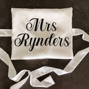 May include: A square white satin pillow featuring the black text Mrs Rynders printed in a cursive font. The pillow has long white satin ribbons attached to the sides for tying, creating a smooth and reflective surface finish on the fabric.