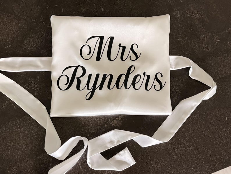 May include: A square white satin pillow featuring the black text Mrs Rynders printed in a cursive font. The pillow has long white satin ribbons attached to the sides for tying, creating a smooth and reflective surface finish on the fabric.