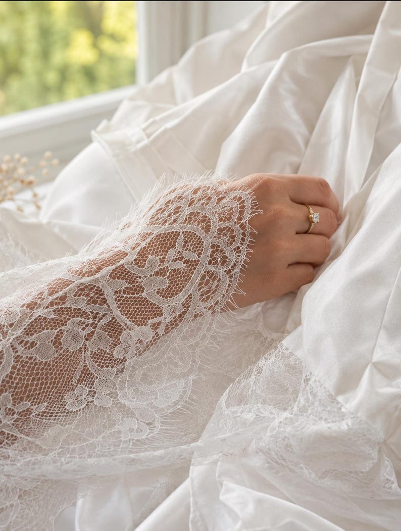 May include: A white long-sleeved garment featuring intricate floral lace detailing on the cuffs and hem, paired with a gold ring set with a single clear stone. The fabric is smooth and opaque, contrasting with the delicate, sheer lace trim.