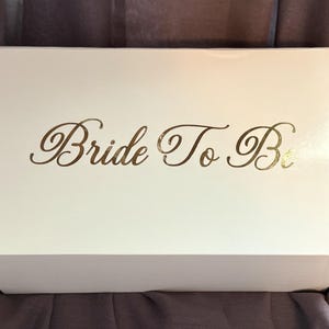 May include: A rectangular white gift box with a smooth finish. The top of the box features the text "Bride To Be" printed in a metallic gold script font. The box is positioned horizontally, showcasing a clean and simple design for storage.
