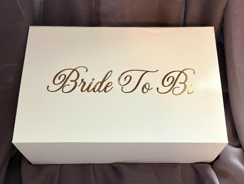 May include: A rectangular white gift box with a smooth finish. The top of the box features the text "Bride To Be" printed in a metallic gold script font. The box is positioned horizontally, showcasing a clean and simple design for storage.